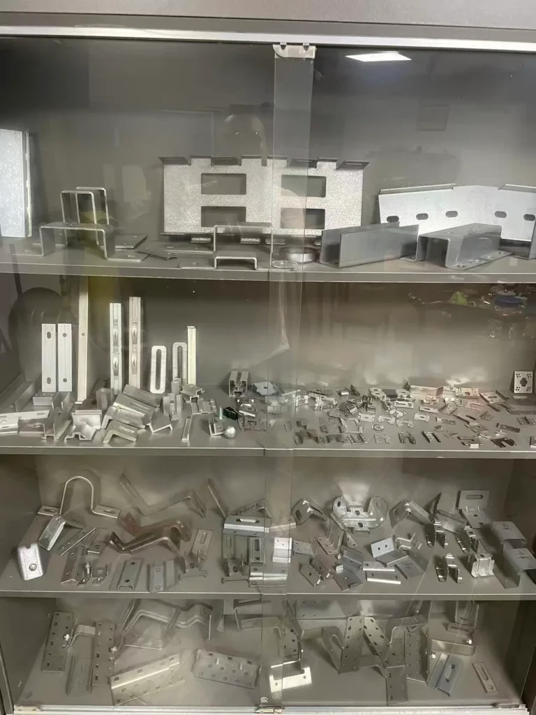 Glass display cabinet with four shelves filled with various custom stamped metal parts for solar products, including brackets, clips, hinges, frames, angle pieces, and structural components.